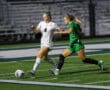 Two female soccer players compete for the ball during a night match on a green field.