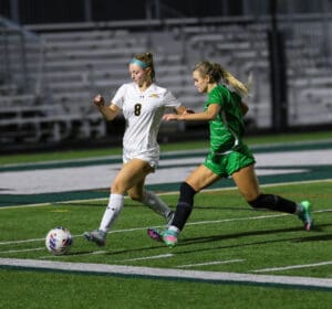 Two female soccer players compete for the ball during a night match on a green field.