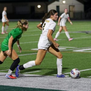 Soccer player in white dribbling the ball, chased by player in green on a field during a night match.