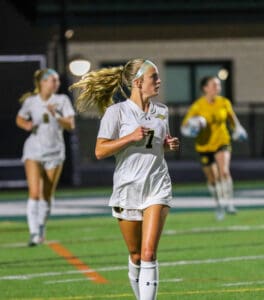 Female soccer player in white uniform runs on field during night game.