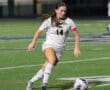 Soccer player in white jersey dribbling during game on turf field at night.
