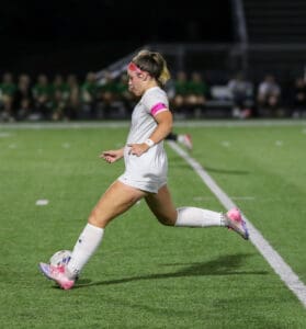 Soccer player in action, kicking ball on field during evening match, wearing white jersey and pink headband.