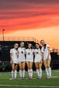 Teen soccer team posing on field at sunset, wearing white uniforms, smiling energetically.