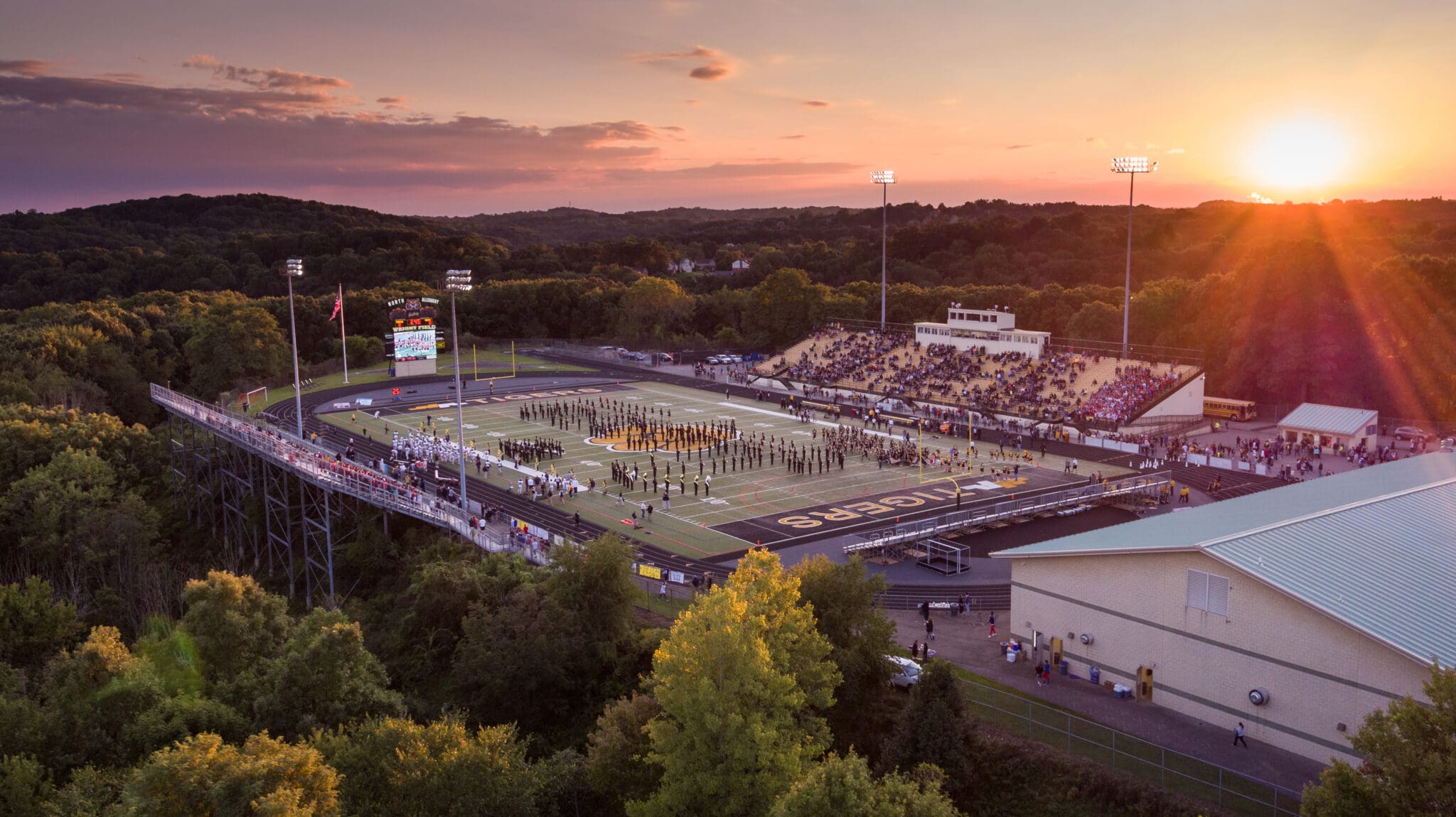 Aerial view of a football stadium with a sunset backdrop, full stands, and marching band on field.