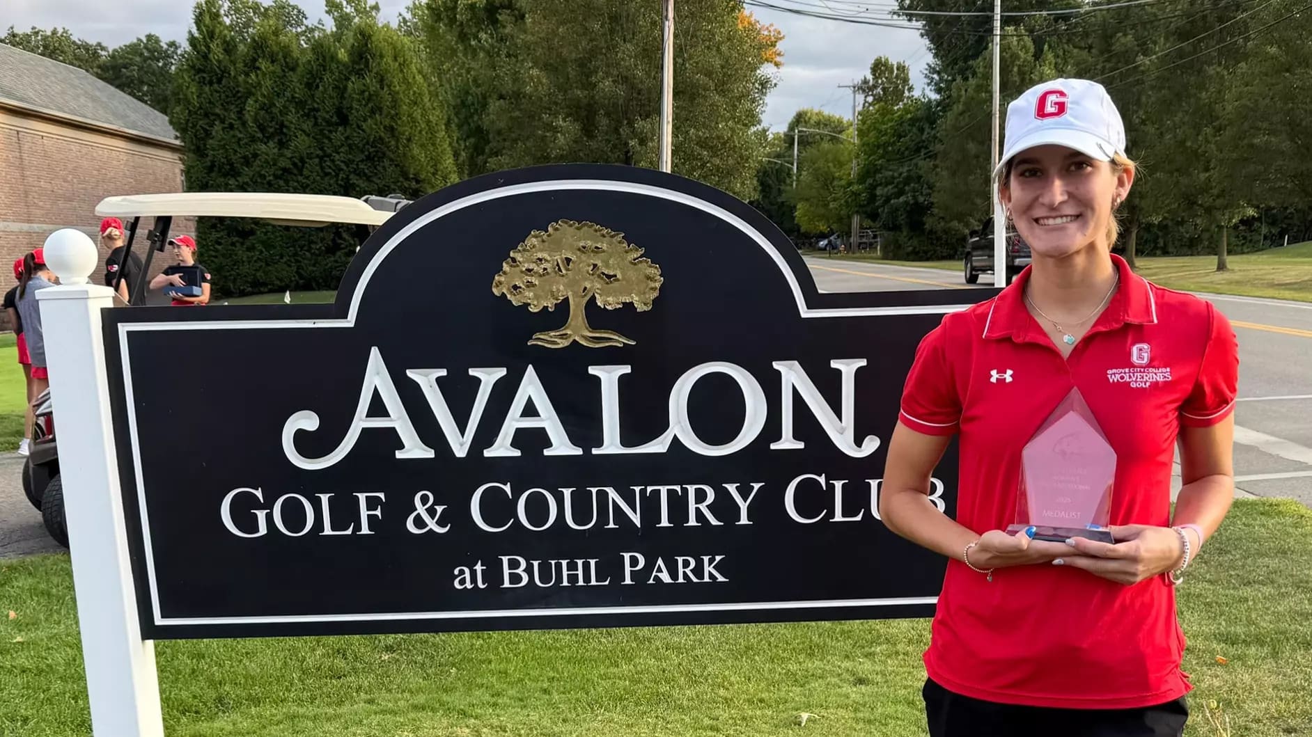 Golfer in red shirt poses with trophy at Avalon Golf & Country Club sign.