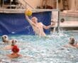 Water polo goalie leaping to block a shot during a competitive match in an indoor pool.