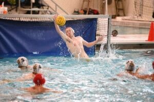 Water polo goalie leaping to block a shot during a competitive match in an indoor pool.