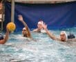 Water polo match with players in blue and white helmets near a goal, players raising arms to block a shot.