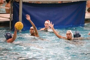 Water polo match with players in blue and white helmets near a goal, players raising arms to block a shot.