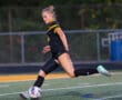 Female soccer player in action, kicking a ball on a field, wearing a black and yellow uniform.