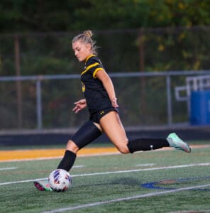 Female soccer player in action, kicking a ball on a field, wearing a black and yellow uniform.