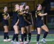 Soccer players in black uniforms celebrate on field after a goal during a night match.