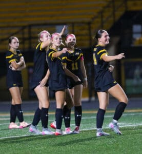 Soccer players in black uniforms celebrate on field after a goal during a night match.
