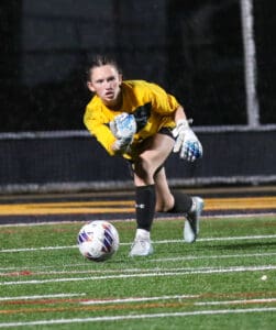 Soccer goalie in yellow saves ball during night game on artificial turf field.