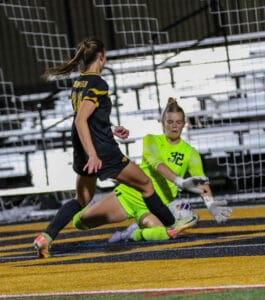 Soccer player in black attempts to score as goalkeeper in green dives to save on a vibrant field.