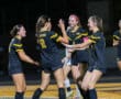 Four female soccer players in black jerseys celebrating on the field at night.