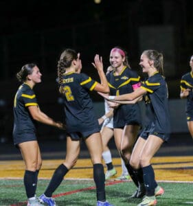 Four female soccer players in black jerseys celebrating on the field at night.