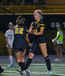 Two female soccer players in black jerseys exchange a high-five during a game on a lit-up field.