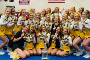 Cheerleading team in yellow uniforms celebrating with a trophy and medals indoors.
