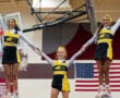 Three cheerleaders in black and yellow uniforms performing a pyramid stunt in a gym with an American flag backdrop.