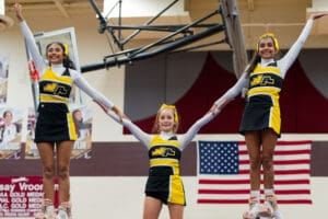 Three cheerleaders in black and yellow uniforms performing a pyramid stunt in a gym with an American flag backdrop.