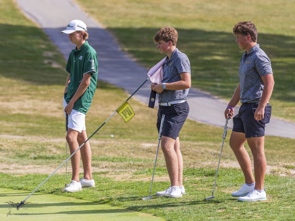 Three young golfers converse on a sunny day at the golf course, standing on the green with their clubs.