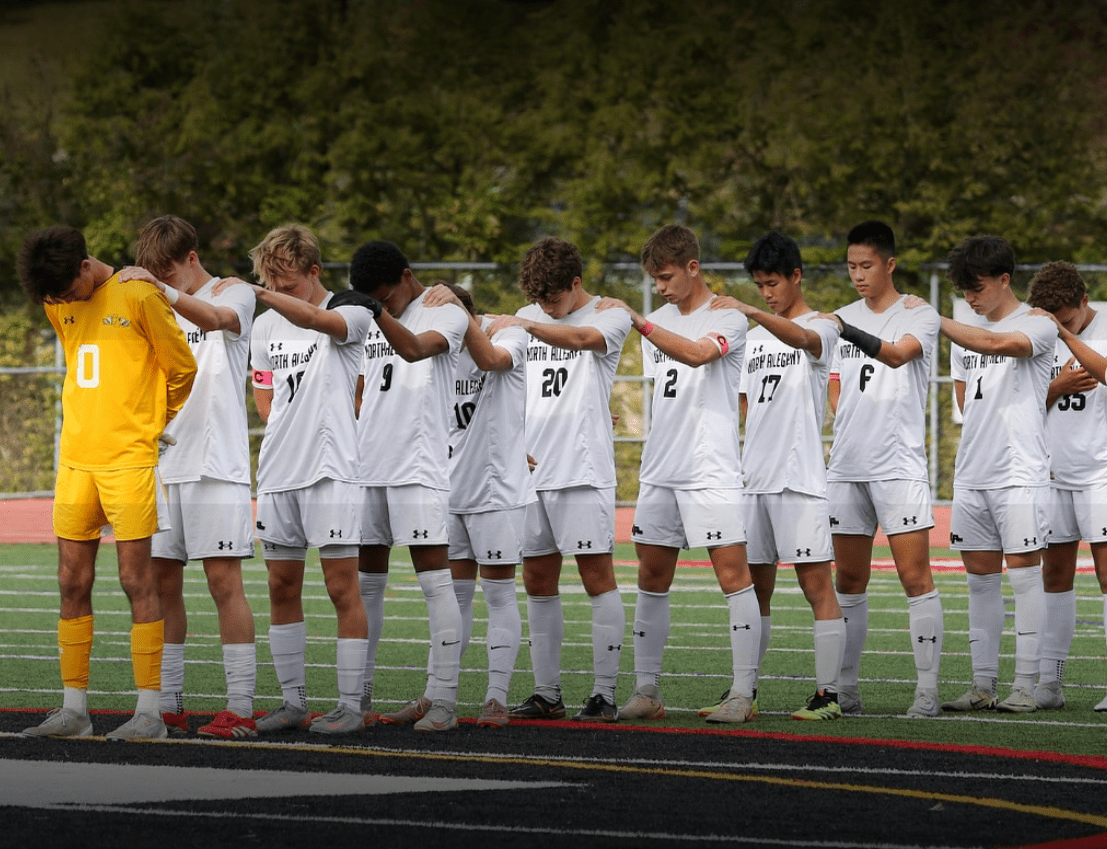 High school soccer team in white jerseys standing together on field, arm in arm, pregame ritual.