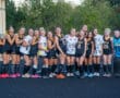 Field hockey team posing together in uniform, smiling at the camera with trees in the background.