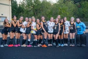Field hockey team posing together in uniform, smiling at the camera with trees in the background.