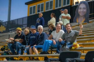 People enjoying a sports event, one holding a large face cutout, seated on stadium bleachers at night.