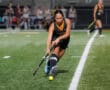 Field hockey player focused on hitting ball during a match at night on green turf.