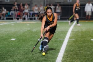 Field hockey player focused on hitting ball during a match at night on green turf.