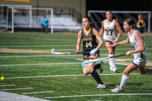 Girls playing field hockey at night, wearing black and white uniforms, chasing the ball on a green field.