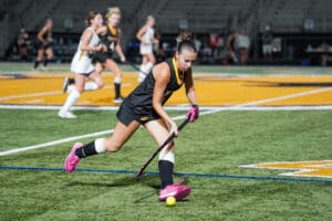 Field hockey player in black uniform dribbling the ball on a brightly lit turf field at night.