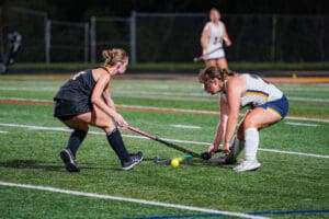 Two female field hockey players compete for the ball on a vibrant green field during a night match.