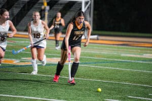 Girls playing field hockey on turf, player in black uniform dribbling the ball under stadium lights.