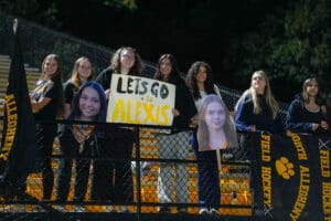 Fans cheering with Let's Go Alexis sign at a sports event, holding banners and cutout faces, nighttime setting.