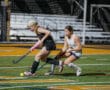 Two female field hockey players competing for the ball on a brightly lit turf field.