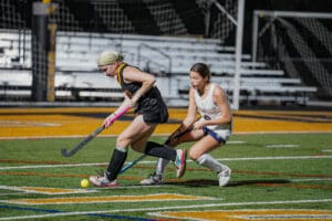 Two female field hockey players competing for the ball on a brightly lit turf field.