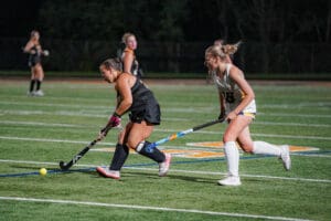 Two field hockey players competing for the ball during a nighttime match on a grassy field.