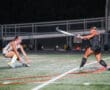 Two field hockey players in action on a night game, one striking the ball, the other defending.