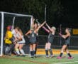 Field hockey team celebrates a goal at night, players in black and goalie in yellow gear.