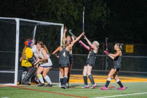Field hockey team celebrates a goal at night, players in black and goalie in yellow gear.