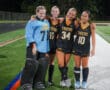 Four field hockey players in team uniforms posing on the field after a game, smiling and embracing.