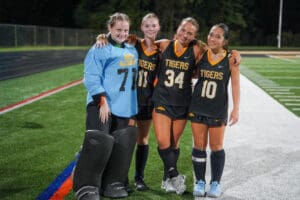 Four field hockey players in team uniforms posing on the field after a game, smiling and embracing.