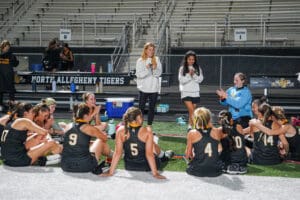 Field hockey team huddles on the turf, listening and clapping, next to North Allegheny Tigers banner.