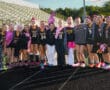 Girls' field hockey team poses on track in uniform with pink accessories and props for themed event.