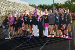 Girls' field hockey team poses on track in uniform with pink accessories and props for themed event.