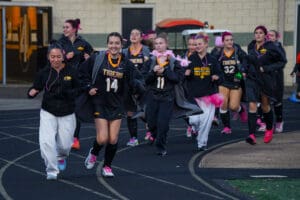 Field hockey team in black uniforms jogging on track, smiling and wearing themed accessories.