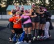 Field hockey team in a group huddle, wearing pink and black uniforms with bows, showing team spirit.